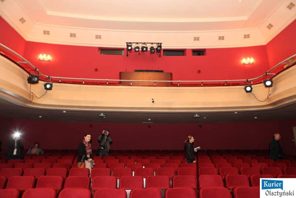 Widok odrestaurowanej sali teatralnej z czerwonymi ścianami, fotelami i oświetleniem na scenie.
