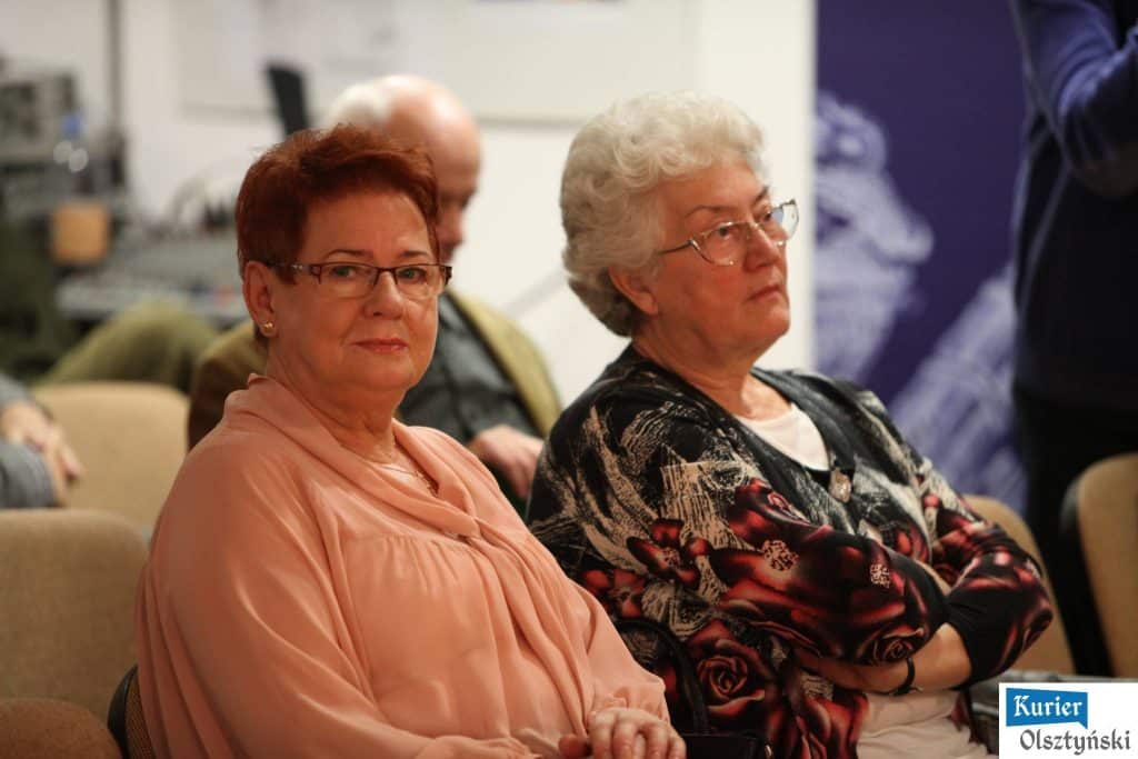 Dwie kobiety siedzą na sali podczas uroczystości, jedna z nich ma okulary i jasny wierzch, druga ma krótkie siwe włosy i czarny sweter z czerwonymi różami.