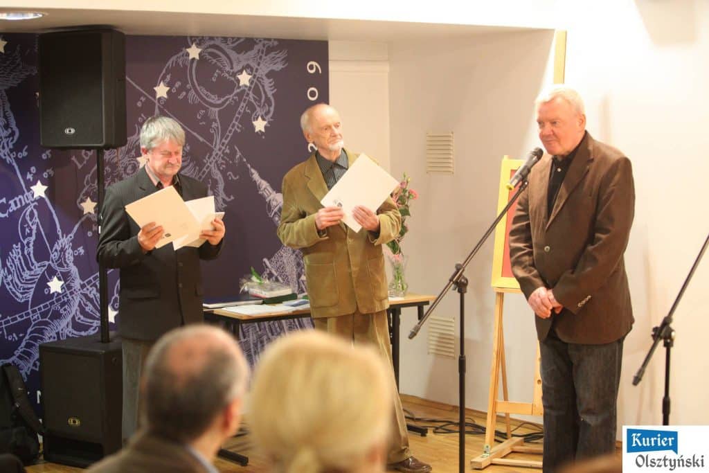 Trzech mężczyzn na scenie podczas ceremonii rozdania nagród, jeden z nich mówi do mikrofonu.