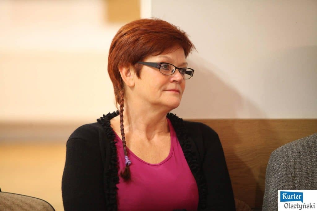Kobieta z czerwonymi włosami, nosząca okulary i różową bluzkę, siedzi na tle jasnej ściany.