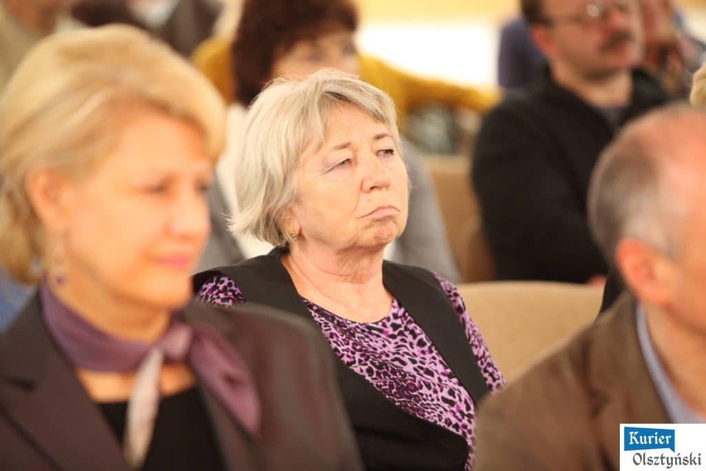 Siedząca kobieta w kwiecistej bluzce na tle innych osób w sali, podczas wydarzenia.