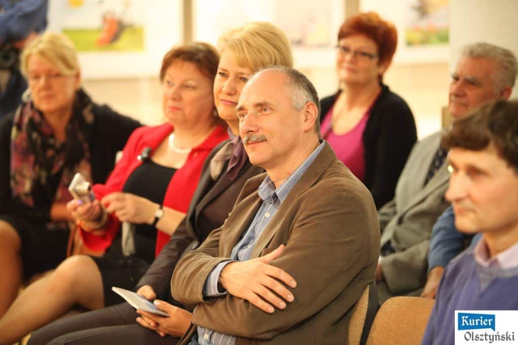 Grupa ludzi siedzi na widowni podczas ceremonii wręczenia nagród, wśród nich mężczyzna w brązowej kurtce i niebieskiej koszuli.