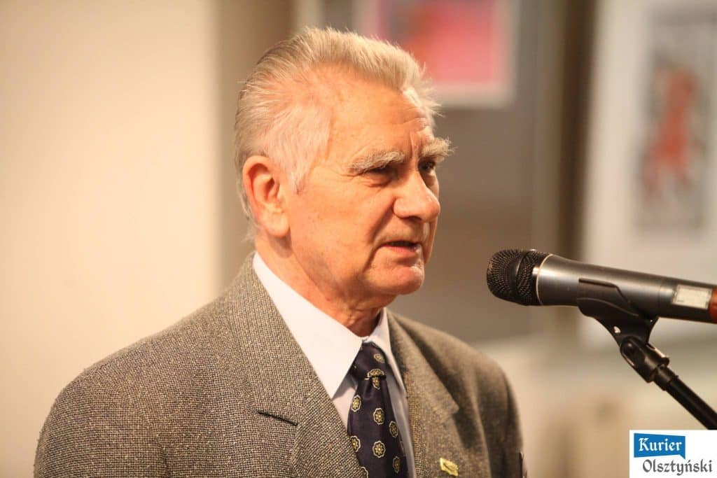 Starszy mężczyzna w garniturze przemawia do mikrofonu podczas wydarzenia, za nim widoczne plakaty.