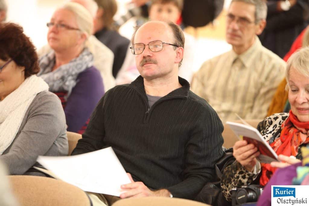 Mężczyzna z brązowym włosem, okularami i w czarnej swetrze siedzi na widowni.