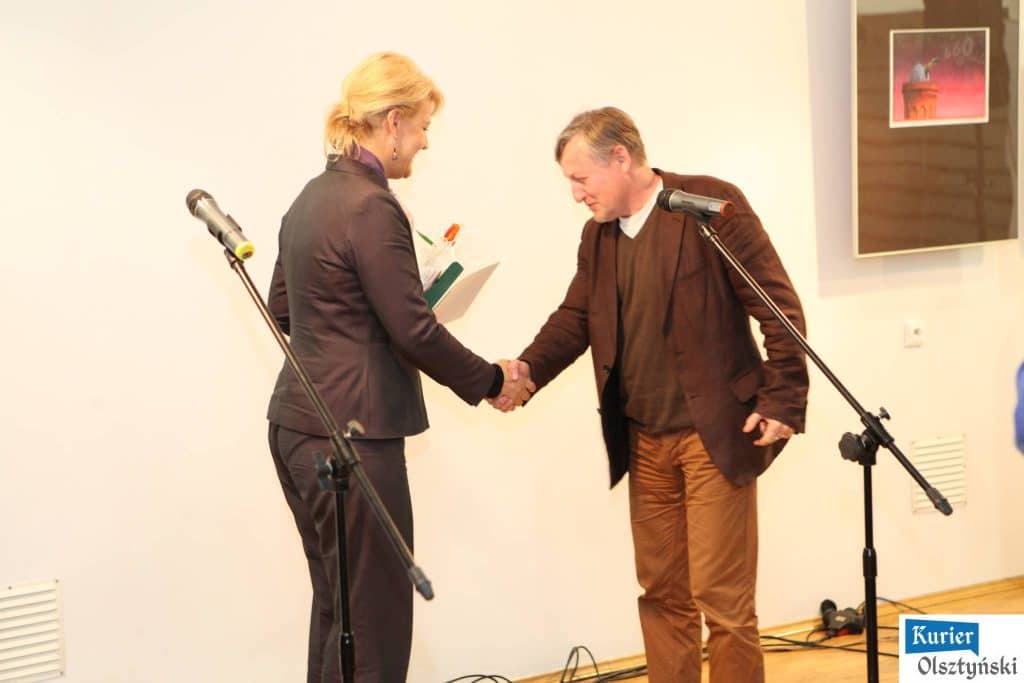 Osoba wręczająca nagrodę satyrykowi podczas ceremonii podczas konferencji, obie osoby uśmiechnięte i witające się.