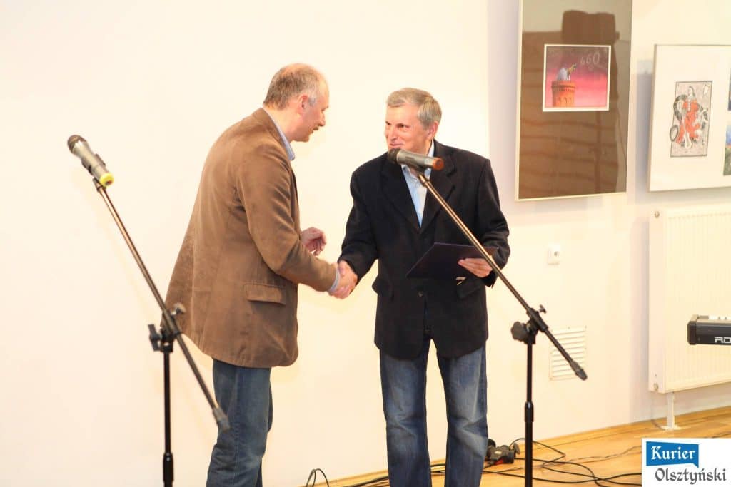 Dwoje mężczyzn uśmiecha się i ściska ręce podczas ceremonii wręczenia nagród, w tle obrazy na ścianie.