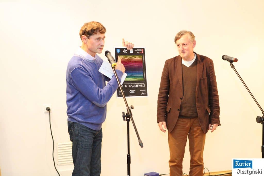 Dwa mężczyźni na scenie podczas wręczania nagród w konkursie satyrycznym, jeden z nich trzyma plakat z kolorowym wykresem i mówi do mikrofonu.
