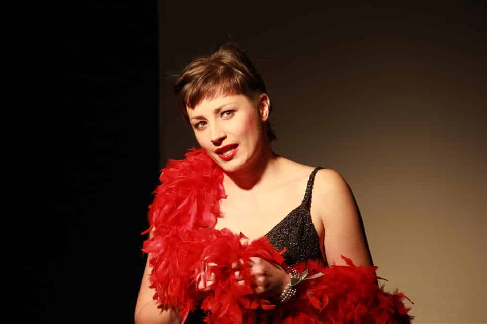 Kobieta na scenie w teatrze, ubrana w czarny strój z czerwonym piórkiem, o krótkich włosach, podczas występu.