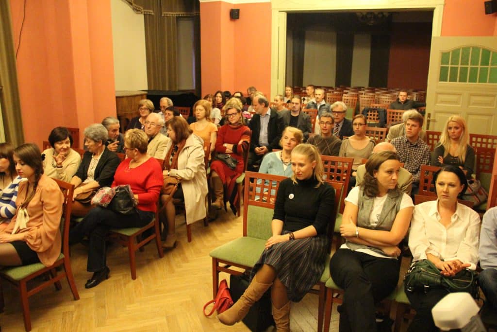 Liczny zespół słuchaczy podczas wydarzenia w Kamienicy Naujackiej, siedzący na drewnianych krzesłach, z widokiem na scenę.