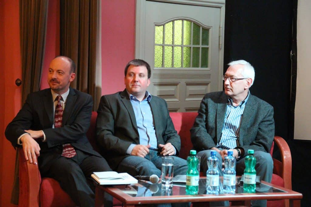 Trzej mężczyźni siedzą na scenie podczas panelu dyskusyjnego, z mikrofonem i notatkami na stole.