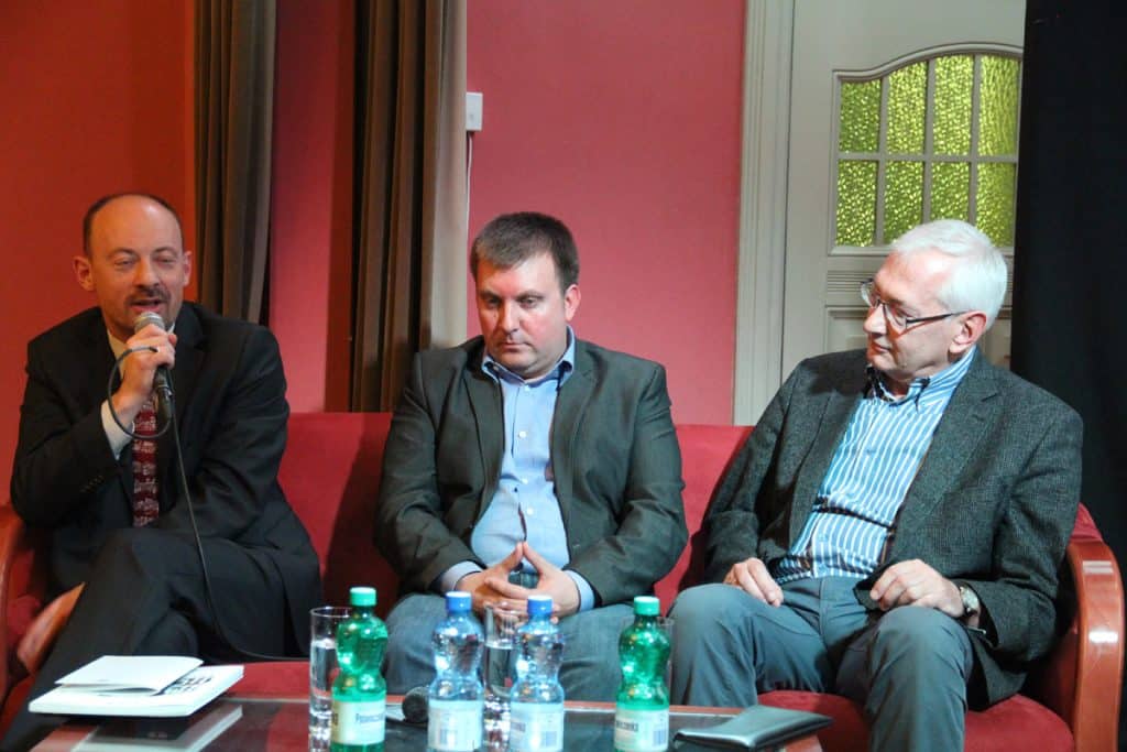 Trzej mężczyźni siedzą na czerwonej sofie podczas dyskusji, w tle okno z zielonym widokiem.