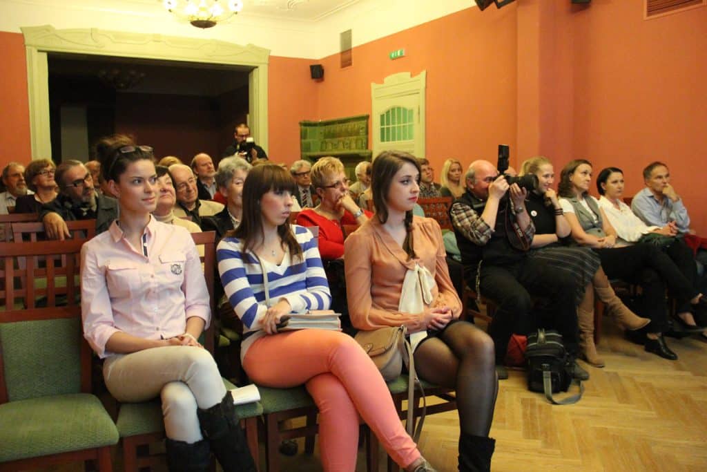 Uczestnicy i goście na wydarzeniu kulturalnym w Kamienicy Naujackiej, siedzą i słuchają prelekcji.
