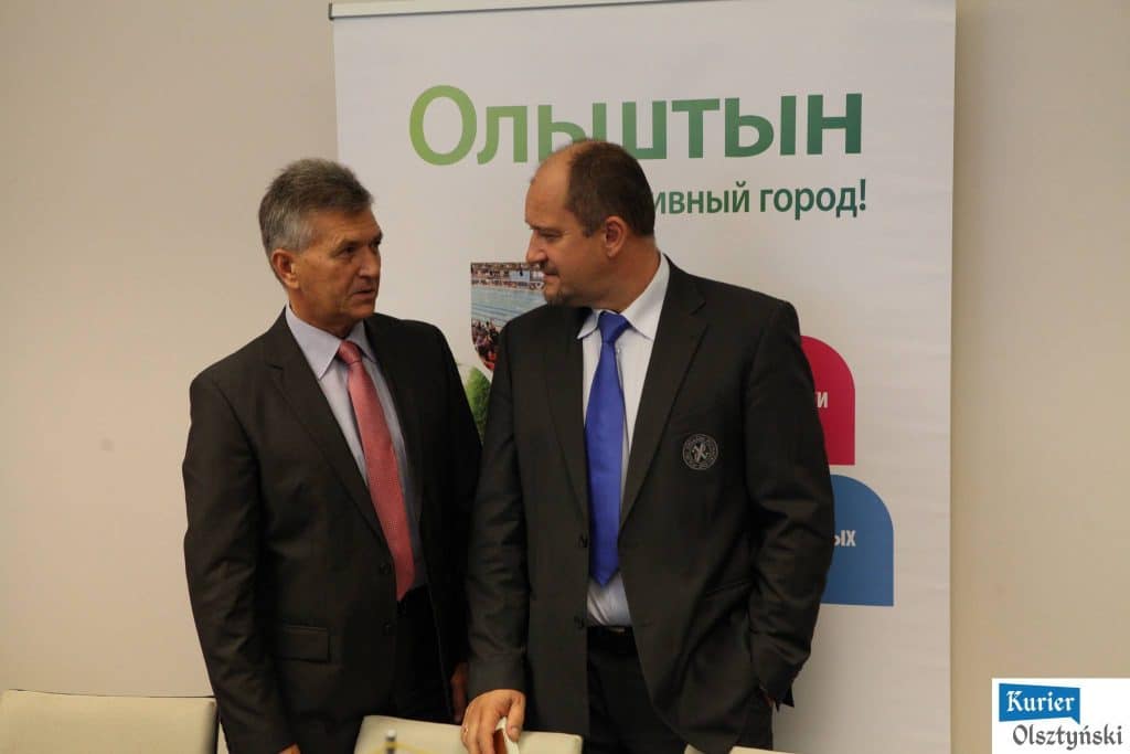 Dwóch mężczyzn w garniturach rozmawia przy stojącej tablicy w pomieszczeniu.