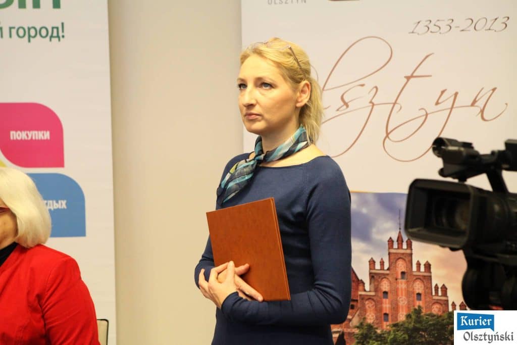 Dorosła kobieta w niebieskiej bluzie i chustce na szyi trzyma książkę podczas konferencji, w tle baner z napisem i obraz zamku.