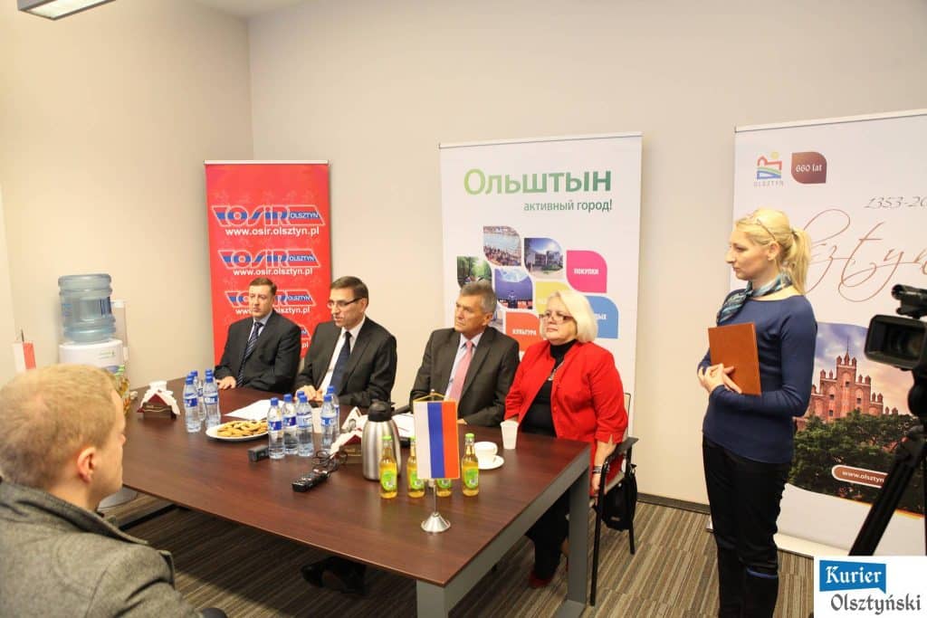 Grupa młodych pływaków z Polski i Rosji podczas konferencji przed zawodami, stoją przy stole z wodą i ciastkami.
