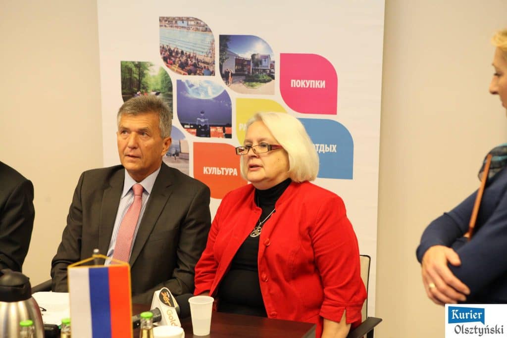 Uczestnicy konferencji przed mitingiem pływackim w Aquasferze, w tym kobieta w czerwonym ubraniu i mężczyzna w garniturze.