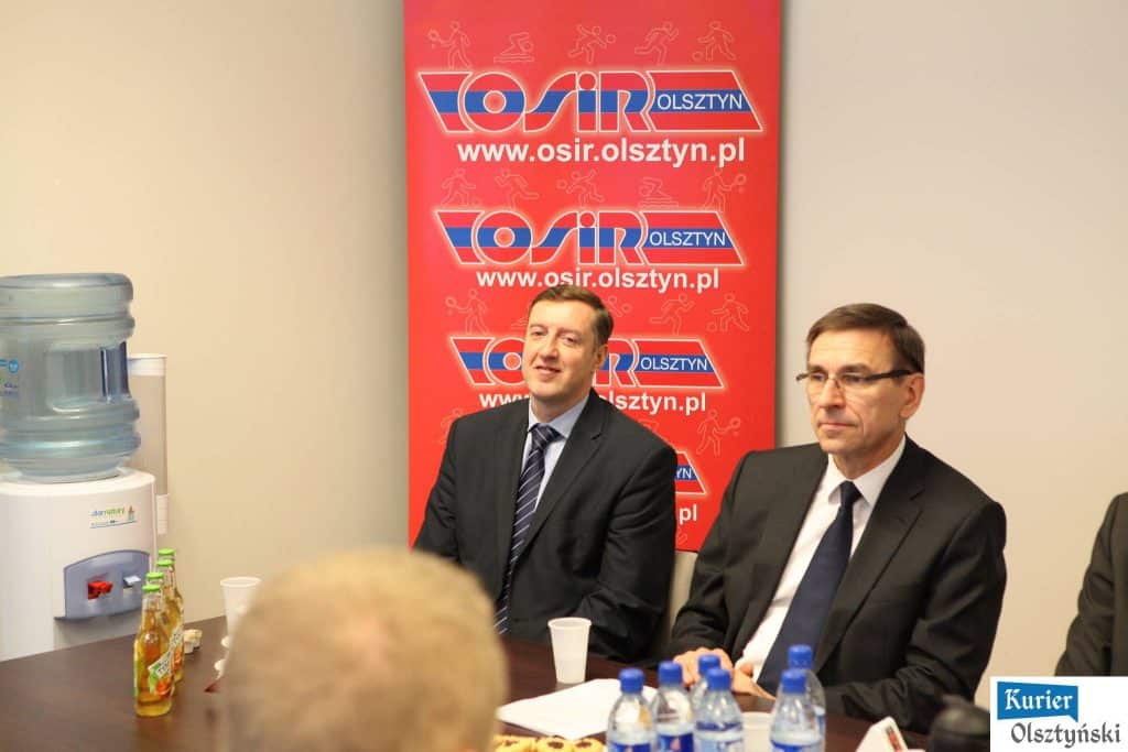 Dwa mężczyźni w garniturowych marynarkach siedzą przy stole podczas konferencji, za nimi czerwony baner z logo osir.