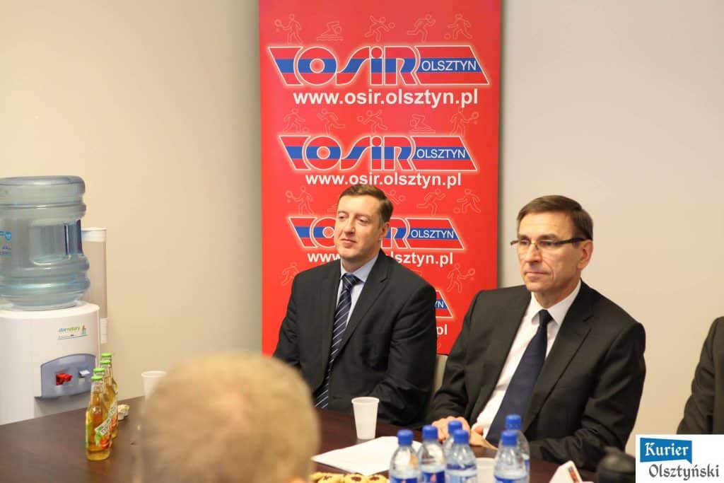 Dwoje mężczyzn w garniturach siedzi przy stole w sali konferencyjnej podczas wydarzenia sportowego.