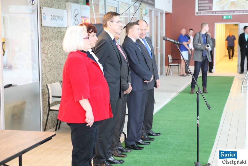 Uczestnicy i organizatorzy mitingu pływackiego w Aquasferze, ustawieni na tle hali sportowej.