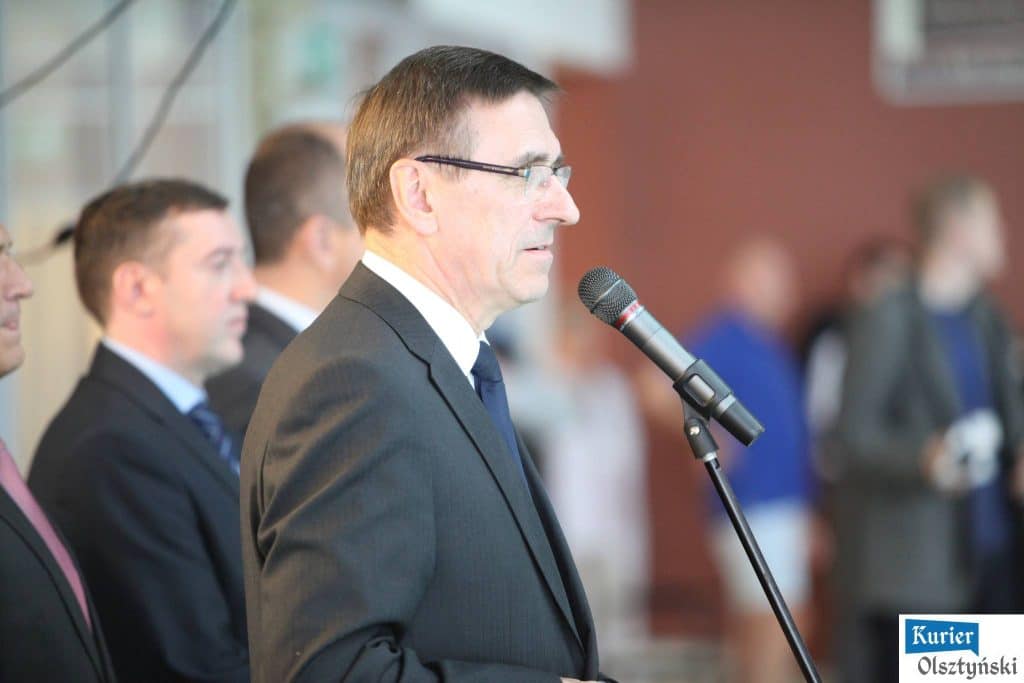 Mężczyzna w garniturze przemawia do mikrofonu podczas wydarzenia, w tle inni uczestnicy.