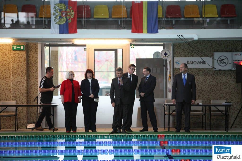 Działacze i sędziowie podczas ceremonii startowej mitingu pływackiego, widoczni na tle torów basenowych i flag krajów.