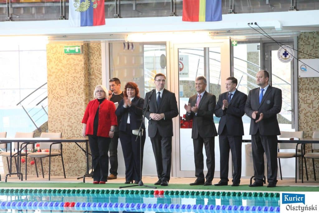 Grupa osób na ceremonii otwarcia mitingu pływackiego w Aquasferze, na tle basenu i flag krajów.