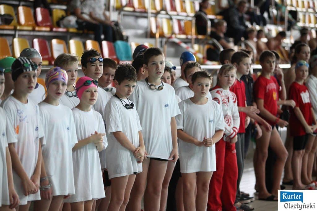 Grupa młodych pływaków w strojach startowych na tle trybun w aquaparku.