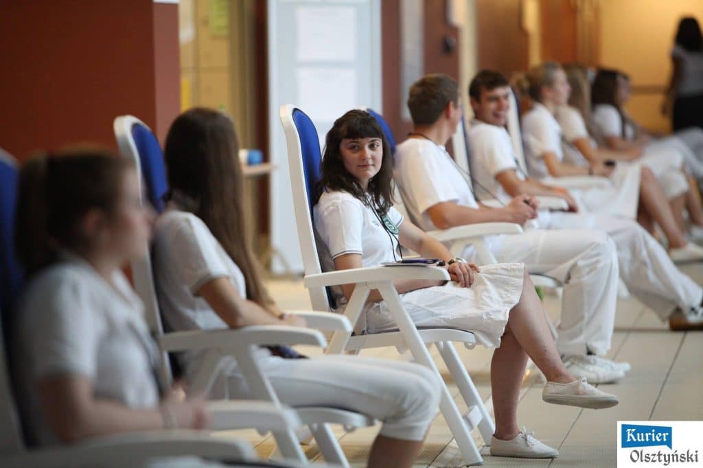 Uczestnicy mitingu pływackiego siedzą w sali, młodzi sportowcy w białych strojach, czekający na start.