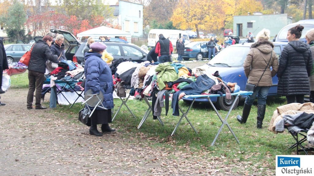 Ludzie na targu z asortymentem odzieży, zabawek i sprzętów na tle samochodów i jesiennego krajobrazu.