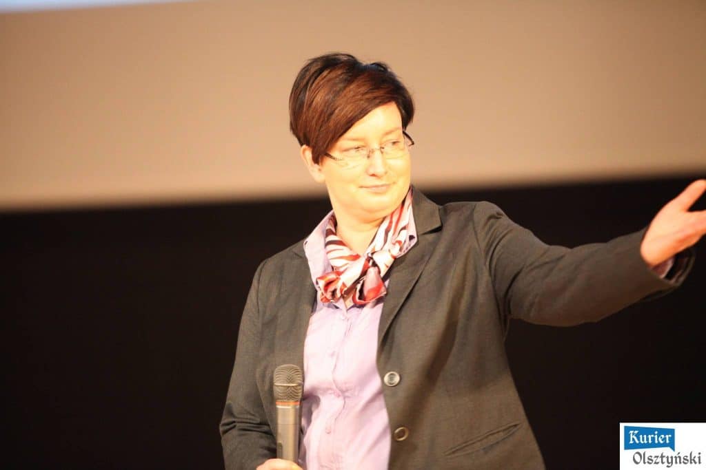 Kobieta w okularach, ubrana w szarą marynarkę i apaszkę, trzyma mikrofon i gestykuluje podczas wydarzenia w sali konferencyjnej.