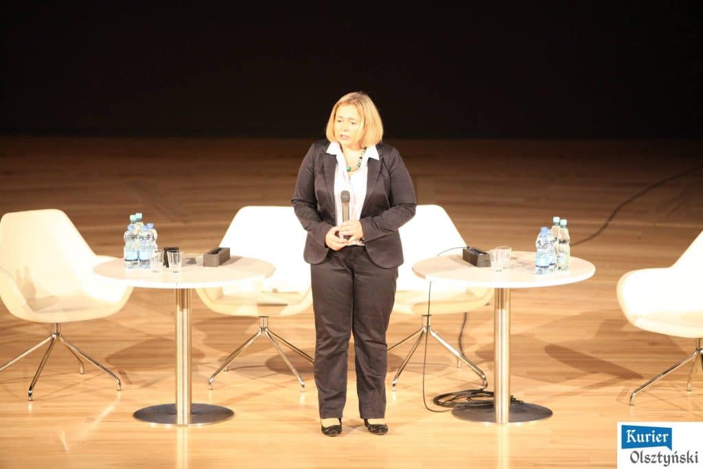 Kobieta z mikrofonem na scenie podczas konferencji w filharmonii, za nią puste krzesła i stoły.