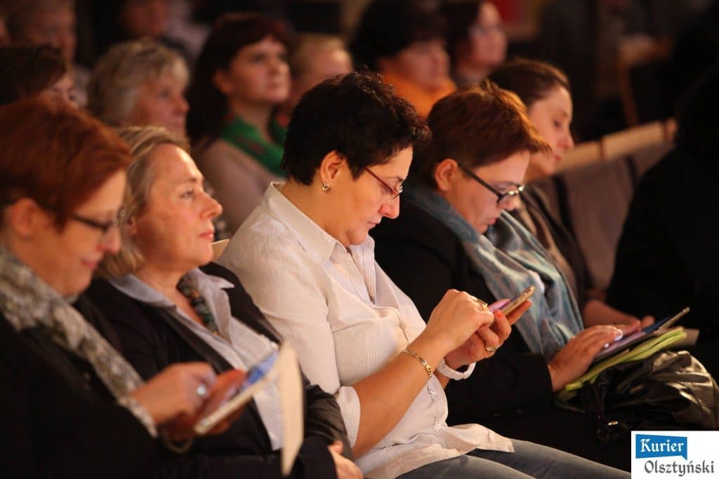 Kobiety siedzą na sali podczas konferencji w filharmonii, korzystają z telefonów i notatek.