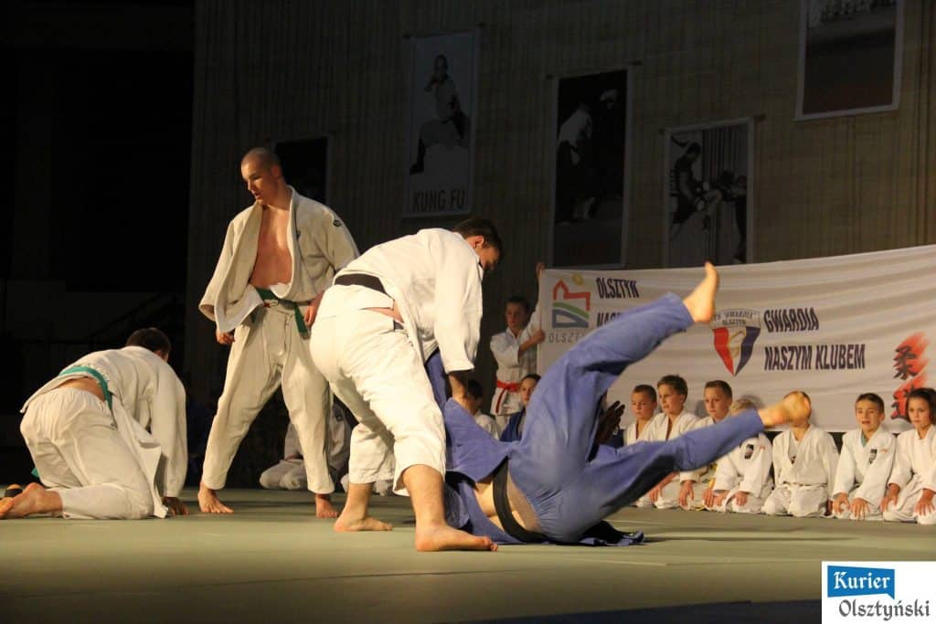 Pokaz sztuk walki na scenie hali Urania w Olsztynie, zawodnicy w judo i tańczący w tle widzowie.