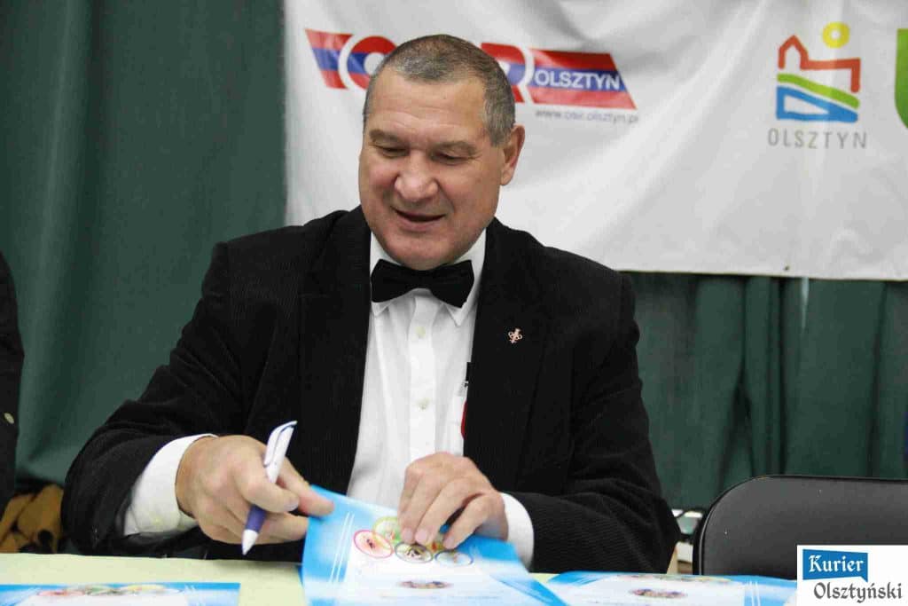Medalista olimpijski i sędzia w ubraniu wieczorowym podpisuje ulotkę, za nim baner z napisem 'Olsztyn' i logo.