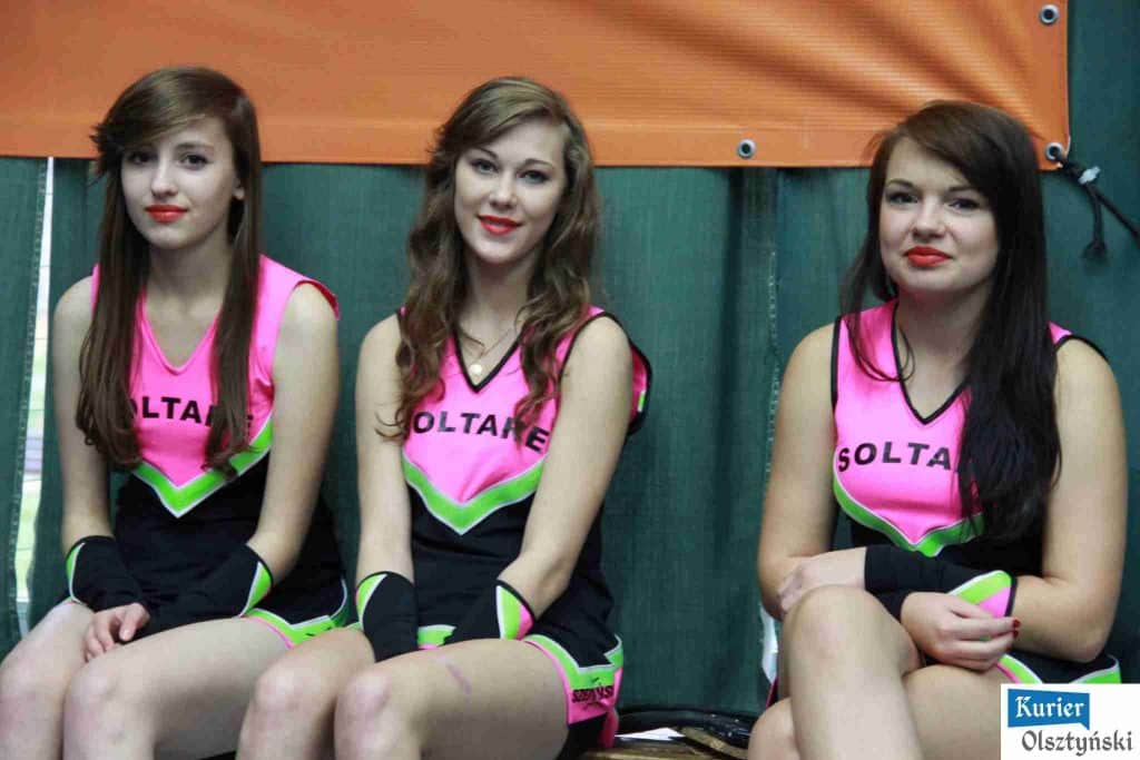 Trzy dziewczyny w różowych strojach cheerleaderek siedzą na ławce podczas wydarzenia sportowego w hali OSiR w Olsztynie.