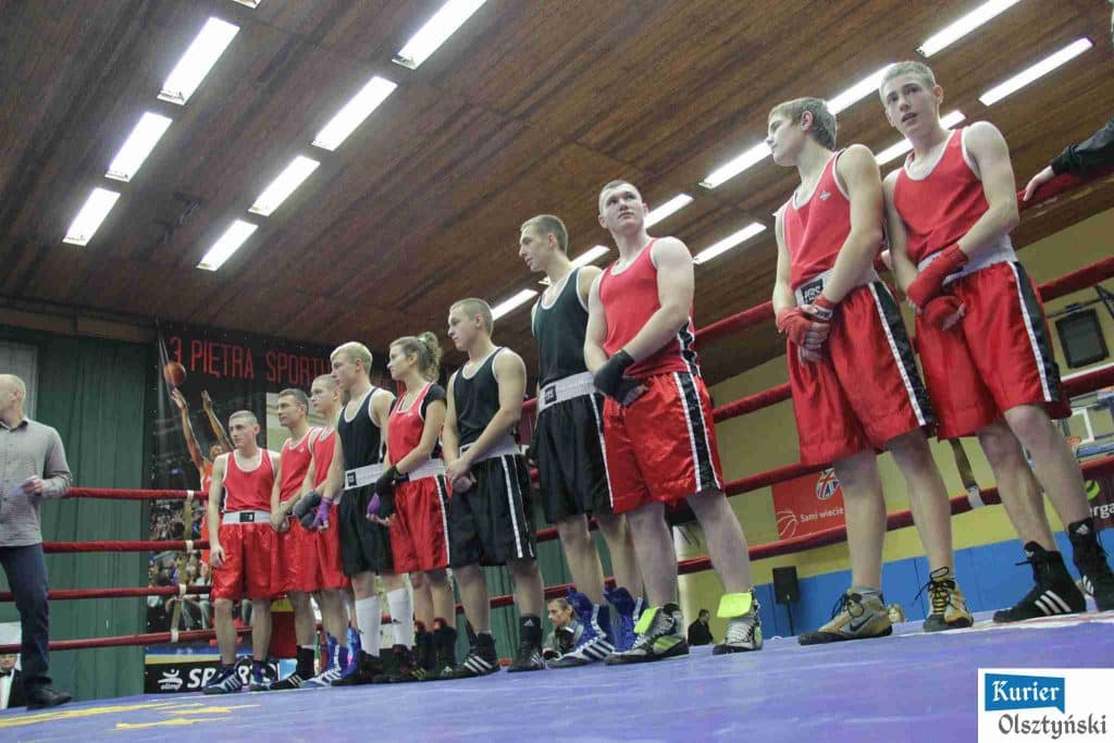 Młodzi bokserzy stojący w ringu podczas wydarzenia sportowego w Olsztynie, ubrane w czerwone i czarne stroje, czekający na rozpoczęcie walki.