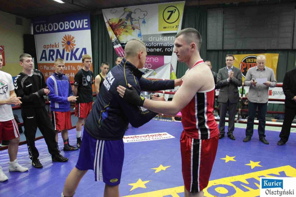 Medalista olimpijski i trener wręczający nagrodę podczas walki w ringu w hali OSiR-u, wokół sędziowie i zawodnicy.