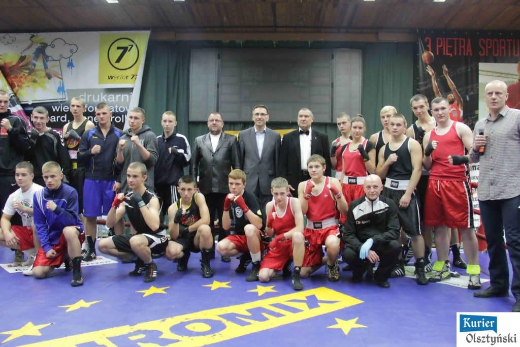 Grupa zawodników i oficjeli na tle ringu podczas wydarzenia sportowego w Olsztynie.