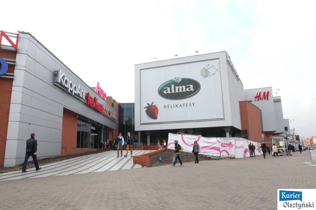 Widok na zewnątrz centrum handlowego Aura z dużym billboardem i osobami spacerującymi po chodniku.