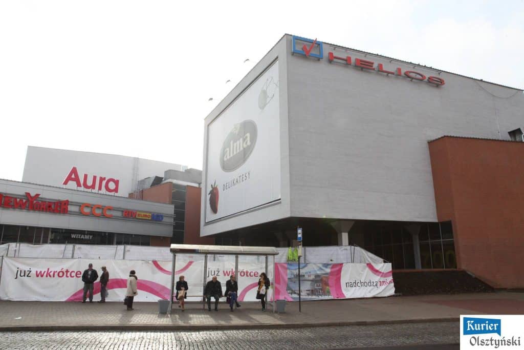 Widok na budynek centrum handlowego Aura z zatłoczonym przystankiem przed wejściem.