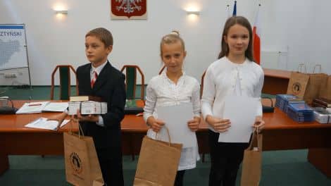 Trzech uczniów stoi na sali, trzymając dyplomy i torby, w tle emblemat i flagi na ścianie.