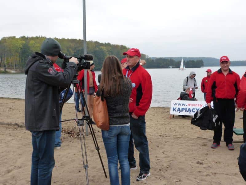 Grupa morsów w strojach przeciwdeszczowych na plaży, ktoś udziela wywiadu, w tle łódka na jeziorze.