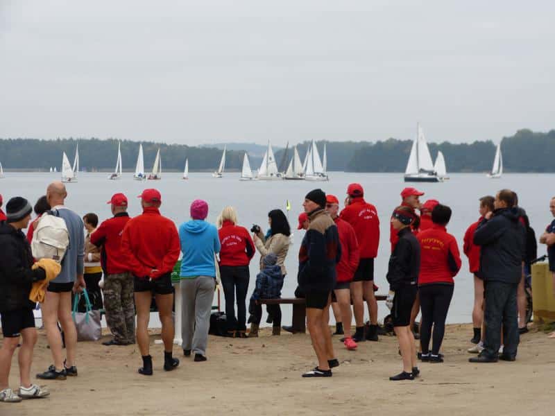 Grupa ludzi na plaży nad jeziorem, część w czerwonych strojach kąpielowych, widok na żaglówki na wodzie.