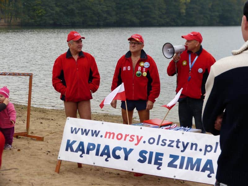 Grupa morsów w czerwonych kurtkach i czapkach na plaży nad jeziorem, z banerem promującym zimowe kąpiele.