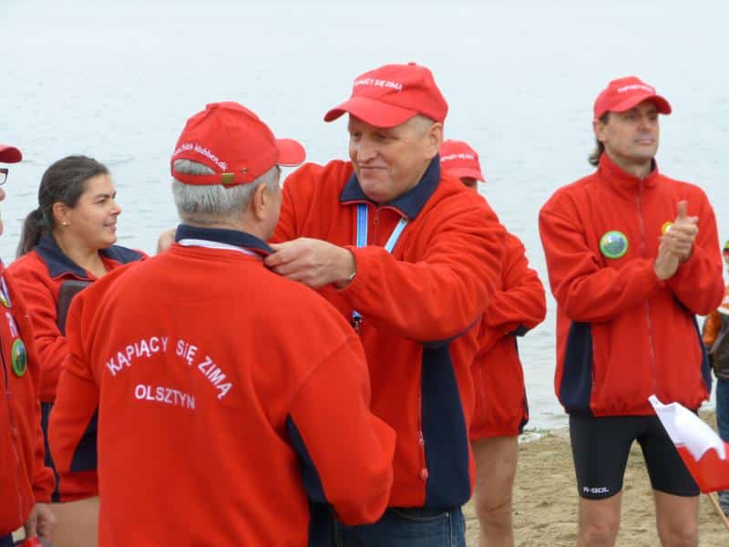 Grupa morsów w czerwonych kurtkach i czapkach podczas inauguracji sezonu na plaży nad jeziorem.