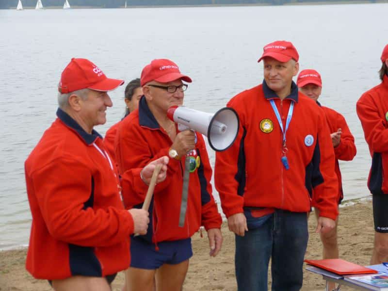 Grupa morsów w czerwonych kurtkach i czapkach, stojąca na brzegu jeziora podczas inauguracji sezonu zimowych kąpieli.