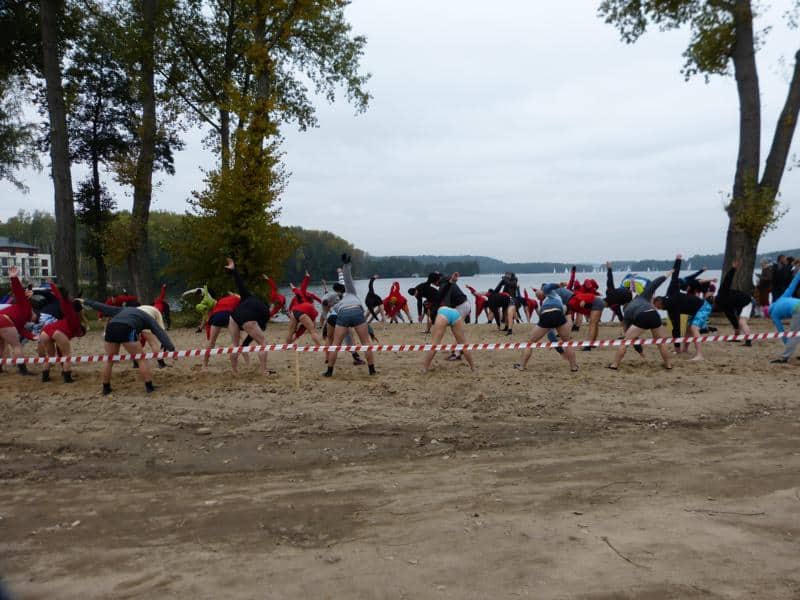 Grupa ludzi na plaży, ubrani w stroje kąpielowe i czapki z uszami, przygotowują się do zimnej kąpieli nad jeziorem.