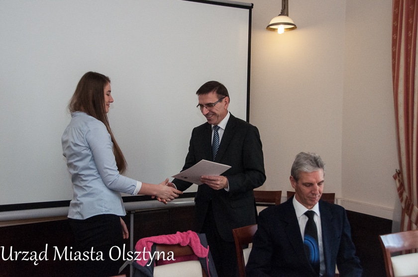 Spotkanie w ratuszu, osoba wręczająca dyplom złotej medalistce taekwondo, około trzech innych uczestników w sali.