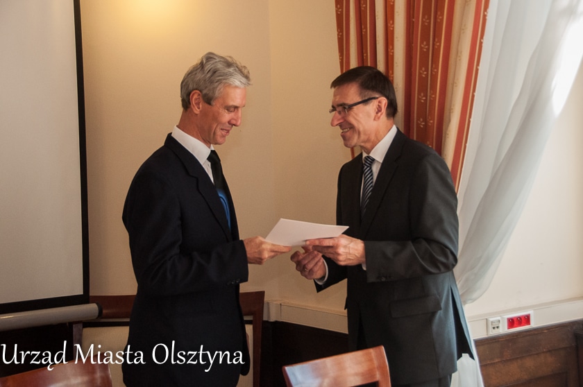 Dwa mężczyźni w oficjalnym spotkaniu, jeden wręcza drugiemu dokument w sali ratusza.