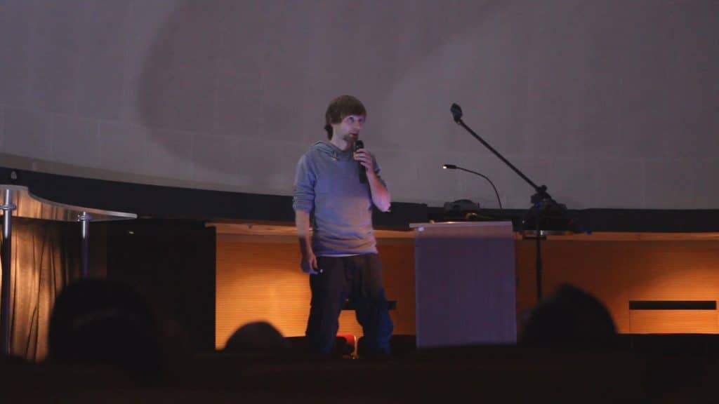 Mężczyzna w bluzie z kapturem mówi do mikrofonu na tle dużego ekranu w sali konferencyjnej.